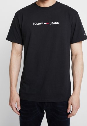 Mężczyzna w czarnej koszulce z krótkim rękawem z białym tekstem "TOMMY JEANS" oraz czerwono-biało-niebieskim logo na piersi, w połączeniu z ciemnymi dżinsami.