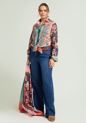 Kvinde iført farverig blomsterprint skjorte med knapper, bundet i taljen, blå jeans, brunt bælte, holder matchende blomsterprint tørklæde, stående mod en ensfarvet baggrund.