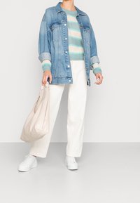 Personne portant une veste en jean bleue, un pull à rayures tricoté, un pantalon blanc, des baskets blanches, tenant un grand sac à main beige.