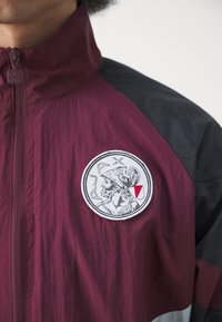 Corta-vento em marrom e preto com um acabamento texturizado, exibindo um patch bordado circular representando uma figura histórica. Detalhe do puxador do fecho visível.