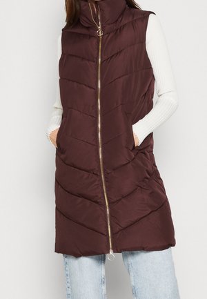 Burgundy padded vest with high collar, zip front, and chevron stitching; features side pockets and a gold zipper. Worn over a white top.