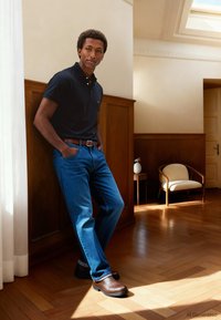 Jūrinės mėlynos spalvos polo marškinėliai, mėlyni džinsai ir rudi batai. Lygus audinys, klasikinio kirpimo, vientisos spalvos. Stovi priešais medinius panelius.