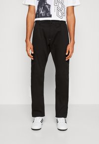 Pantaloni cargo neri a sbieco con tasche laterali, dotati di chiusura con bottone e cerniera. Abbinati a una t-shirt bianca con grafica e sneakers bianche.