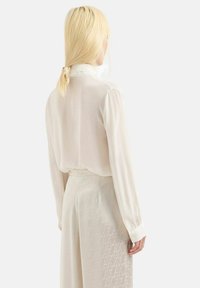 Blusa bianca di seta con maniche lunghe e colletto alto, abbinata a pantaloni leggeri a motivo con un sottile design geometrico.