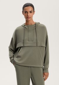 Olivovo zelená mikina s voľným strihom, textúrovaná tkanina, predná vrecko a stiahnuteľná kapucňa.
