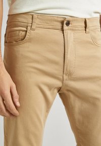 Pantalones de algodón beige con un corte ajustado, con un bolsillo frontal, trabillas para cinturón y un cierre de botón marrón. La tela tiene una textura suave.