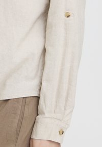 Poignet de chemise en lin beige à manches longues avec manche boutonnée à côté d'un pantalon marron, montrant la texture du tissu et les détails des coutures.
