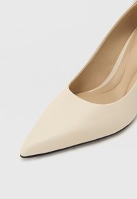 Beige teravatipud jalatsid, millel on sile nahk pealis, madal vöö ja must välistald, näidates minimalistlikku disaini.