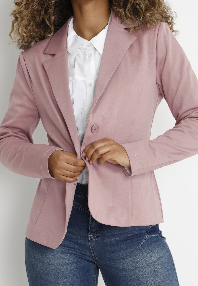 Blazer rosa chiaro in tessuto liscio, con revers a punta, chiusura con un solo bottone e maniche lunghe; indossato sopra una camicia bianca e jeans.