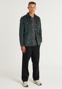 Donkergroen corduroy shirtjack met twee borstzakken, knoopsluiting, gedragen over een grijs T-shirt, gecombineerd met zwarte broek en lichte sneakers.