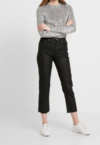 Silberne Pailletten-Bluse mit langen Ärmeln, kombiniert mit hoch taillierten schwarzen Jeans. Die Jeans haben ein gerades Bein und einen rohen Saum. Das Model trägt weiße Sneaker.