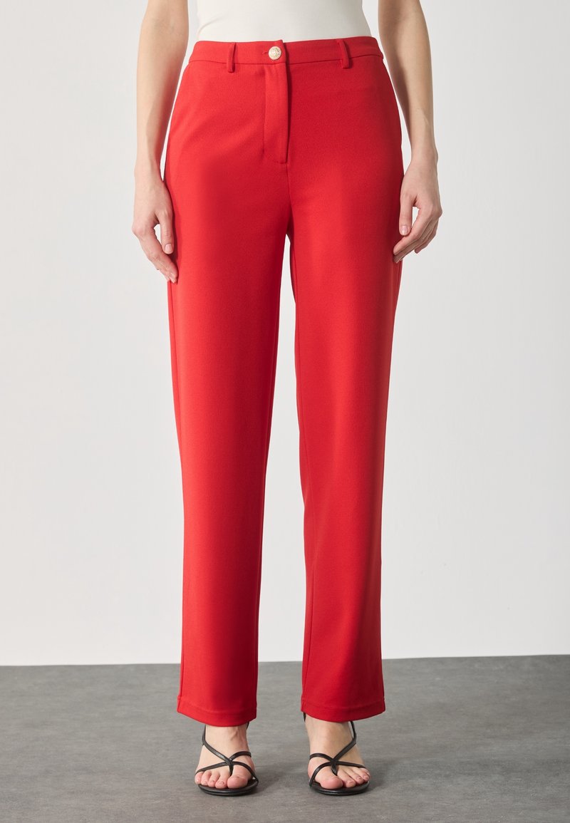 Pantalon rouge droit avec fermeture à bouton et fermeture éclair, porté avec des sandales noires à brides sur un sol gris.