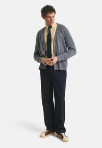 Grauer Cardigan mit Knöpfen, getragen über einem gelben Hemd und einer dunkelgrünen Krawatte mit Punkten. Kombiniert mit dunklen Hosen und lässigen Sneakern.