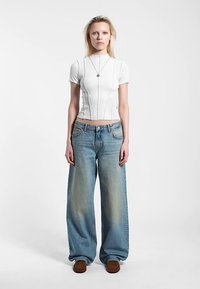 Vit bomullcroptop med vertikala sömmar, kombinerad med ljusblå vida jeans med blekta detaljer och avslappnad passform. Bruna skor.