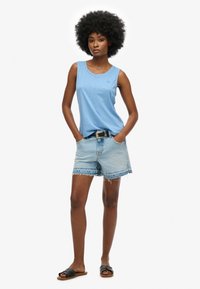 Blauwe mouwloze top, lichte denim shorts met een rafelige zoom en zwarte sliders. Model poseert met handen in de zakken en toont een casual outfit.