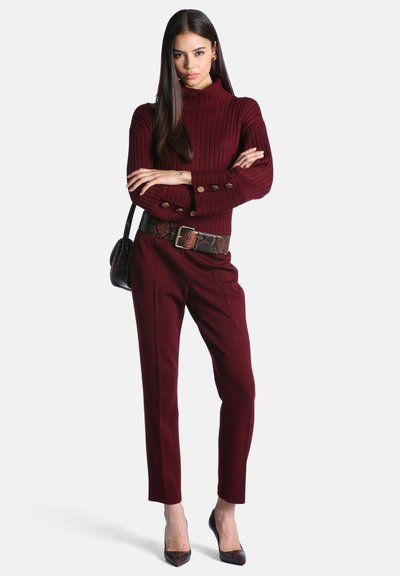 Femme aux longs cheveux foncés portant un pull côtelé bordeaux, un pantalon assorti, une ceinture imprimé serpent et des talons, debout les bras croisés.