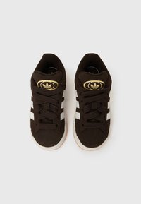 Καφέ σουέντ αθλητικά παπούτσια με λευκές ρίγες και χρυσές λεπτομέρειες λογότυπου, στρογγυλές μύτες και υφασμένα κορδόνια. Η σχεδίαση ολοκληρώνεται με λαστιχένιες σόλες.