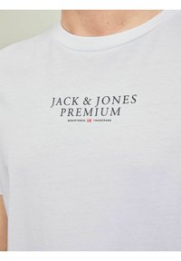 Bílý bavlněný tričko s textem "JACK & JONES PREMIUM" v tučném černém písmu. Kulatý výstřih a krátké rukávy. Jemná, hladká textura.