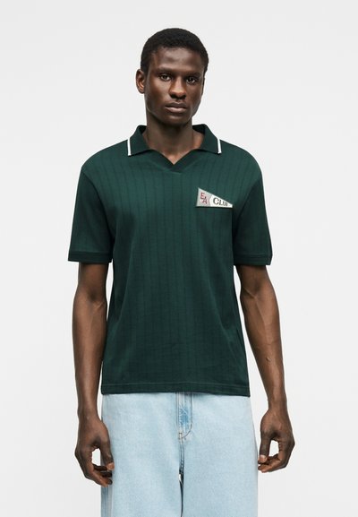 Mężczyzna ubrany w ciemnozieloną koszulkę polo z krótkim rękawem, kołnierzem wykończonym białą lamówką oraz naszywką "EA Club", w połączeniu z jasnoniebieskimi dżinsami.