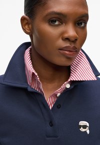 Marinblå polo t-shirt med en randig krage i rött och vitt, med en knappslå och en broderad logotyp av en person med solglasögon.