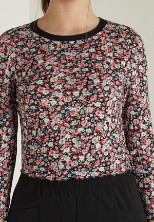 Femme portant un haut noir ajusté à manches longues recouvert d'un motif floral dense rose, blanc et violet, associé à un pantalon noir.