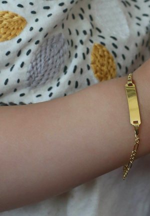 Gouden armband met een rechthoekige bedel, voorzien van een schakeldesign. Draagt op een lichte huidarm met een achtergrond van geweven stof.