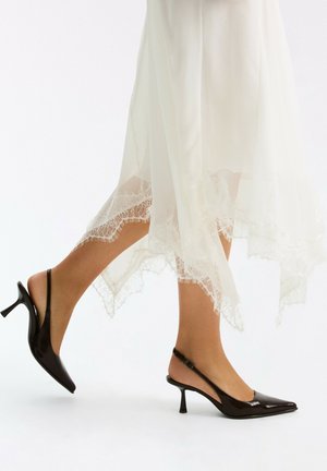 Kvinde iført sorte slingback hæle og en hvid midinederdel med blondekant, går med den ene fod let løftet mod en hvid baggrund.