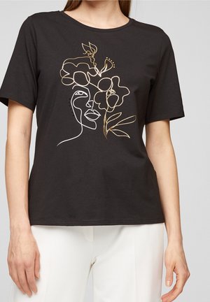 Zwart T-shirt met korte mouwen met een doorlopende lijntekening in wit en goud van een gezicht verweven met bloemen, gedragen met een witte broek.