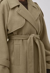 Manteau trench en mélange de laine beige avec grandes revers, taille cintrée et poches latérales. Tissu texturé avec une coupe ajustée et un design classique.