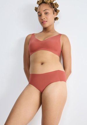 Vrouw met natuurlijk haar in twists, gekleed in een bijpassende naadloze roestkleurige bh en slips tegen een eenvoudige lichte achtergrond.