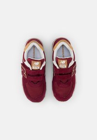 Sneaker marroni con tomaia in suede e rete, dotate di una fibbia in velcro, suola bianca e dettagli dorati. Numero di modello 574 visibile sulla linguetta.