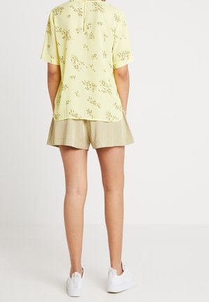 Personne portant un haut à manches courtes floral jaune pâle, un short beige et des baskets blanches, debout avec les mains dans les poches, tournée de dos.