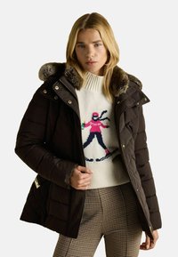 Hnedá pufovaná bunda s kapucňou podšitú kožušinou, so zipsom vpredu a prešívaným dizajnom. Nosená cez pletený krémový sveter s grafikou lyžiara.