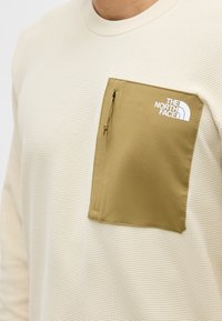 Μπεζ μακρυμάνικο πουκάμισο με υφή, με ταν εξωτερική τσέπη στο στήθος με φερμουάρ και λευκό λογότυπο The North Face στην αριστερή πλευρά.