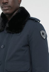 Marineblauwe jas met een zwart nepbonten kraag, voorzien van knopen, een borstpatch en een geweven stofafwerking, ontworpen voor warmte.