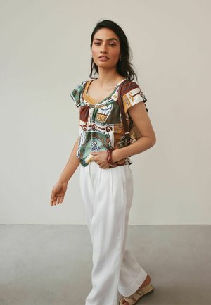 Femeie purtând o bluză cu mâneci scurte cu model, pantaloni albi cu picioare largi, sandale și având o geantă maro pe umăr, stând pe un fundal deschis.