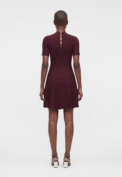 Personne portant une robe rouge foncé à manches courtes, côtelée, avec trois fermoirs dorés sur le haut du dos, debout, dos tourné vers l'avant.