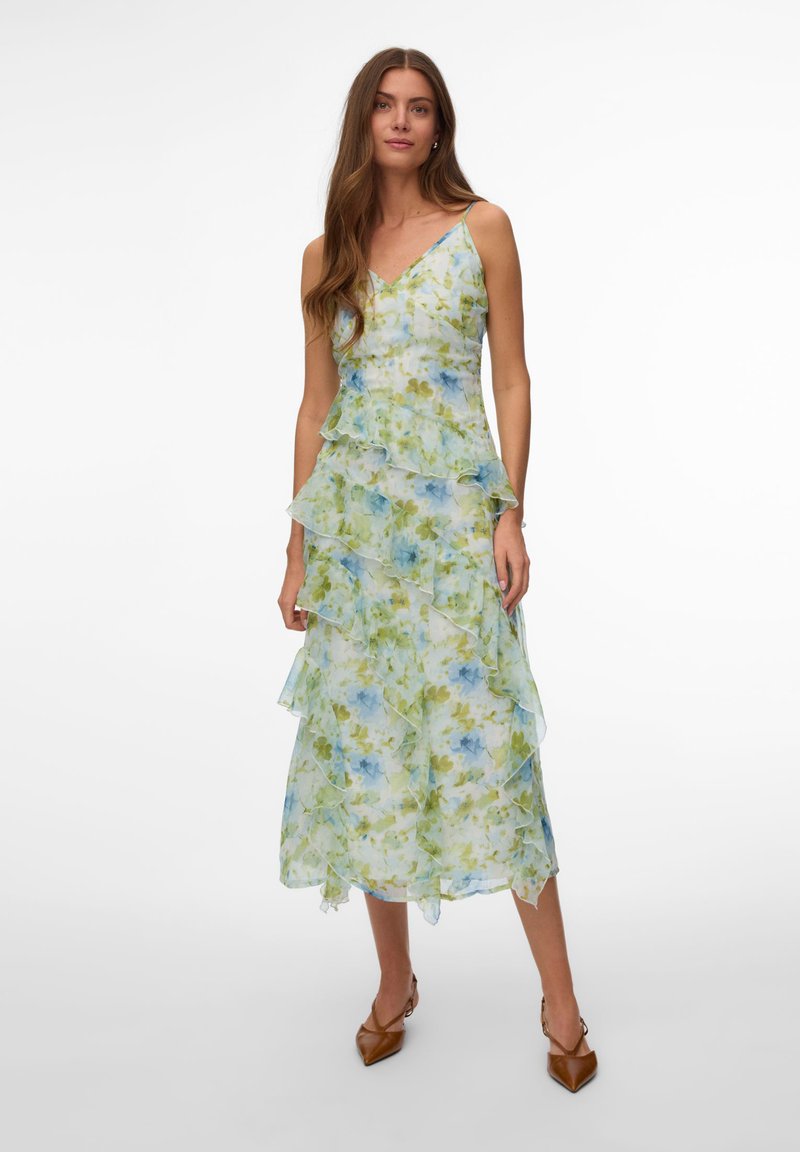 Γυναίκα που φορεί αμάνικο, μέσης μήκους φόρεμα με πράσινο και μπλε λουλουδάτο σχέδιο, σε συνδυασμό με καφέ μυτερές γόβες, στέκεται μπροστά σε λευκό φόντο.