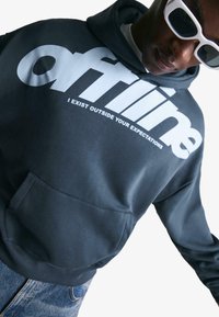 Man wearing dark hoodie with "offline I EXIST OUTSIDE YOUR EXPECTATIONS" text, white sunglasses, and blue jeans against a white background.