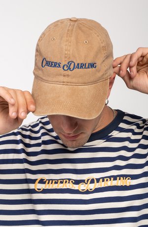 Gorra de béisbol de algodón marrón con texto bordado azul "Cheers, Darling" en la parte frontal, usada ligeramente inclinada. Camisa a rayas azul y blanca debajo.