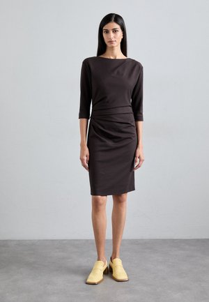 Brown, fitted dress with three-quarter sleeves, gathered waist detail, and smooth texture. Worn with light yellow, woven block-heeled shoes.