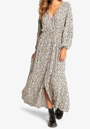 Femme portant une longue robe fleurie à manches longues avec une fente à l'avant, associée à des bottines noires à lacets.