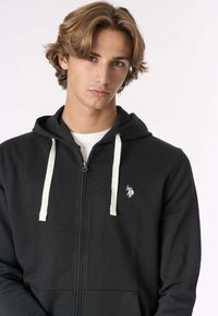 Felpa nera con zip, coulisse bianca, tessuto texturizzato, tasche frontali e piccolo logo sul petto a sinistra.