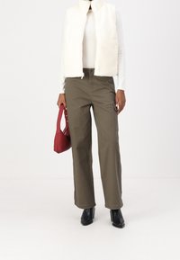 Faux-Fur-Weste in Creme, weißes Langarmoberteil, olivefarbene weit geschnittene Hose, rote Handtasche und schwarze Stiefeletten mit Absatz. Vertikaler Reißverschluss an der Weste.