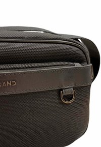 Sac bandoulière noir en tissu avec une surface texturée, des accents en cuir, une fermeture éclair et un détail en anneau en métal circulaire. Logo visible sur le devant.