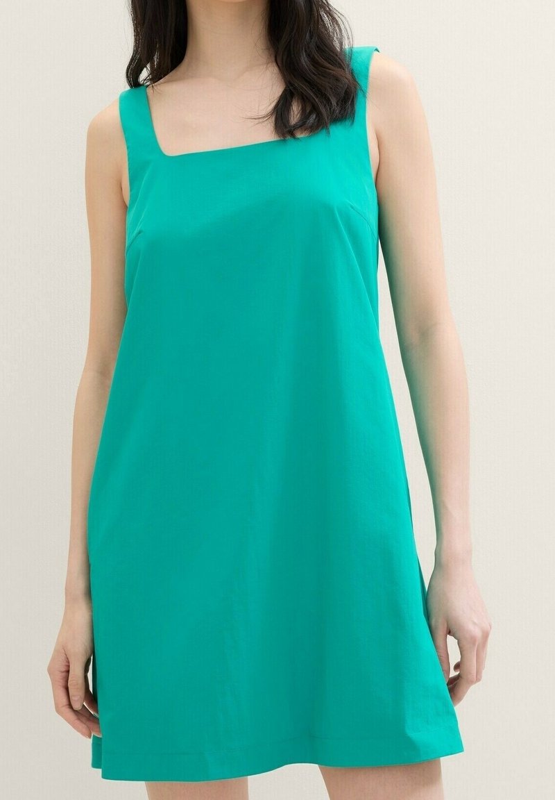 Femme portant une mini robe turquoise sans manches avec un décolleté carré, debout devant un fond clair uni.