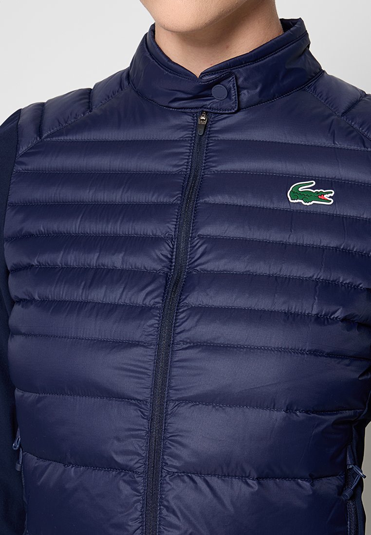 Gilet matelassé bleu marine avec des coutures horizontales, un col haut et une fermeture à glissière complète. Présente un logo crocodile vert distinctif sur la poitrine gauche.