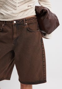 Shorts de mezclilla marrones con textura descolorida, cinco bolsillos y herrajes metálicos; llevados con un bolso de cuero marrón oscuro arrugado.