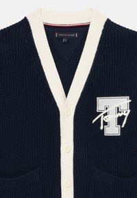 Námořnický pletený cardigan s krémovým lemem, výstřihem do V, pěti knoflíky, dvěma kapsami a vyšitým logem "Tommy" na levé části hrudi.