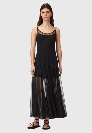 Frau mit langen dunklen Haaren, die ein schwarzes ärmelloses Kleid mit transparentem Mesh-Überzug und schwarze, flache, geraffte Sandalen trägt, steht vor einem einfarbigen Hintergrund.